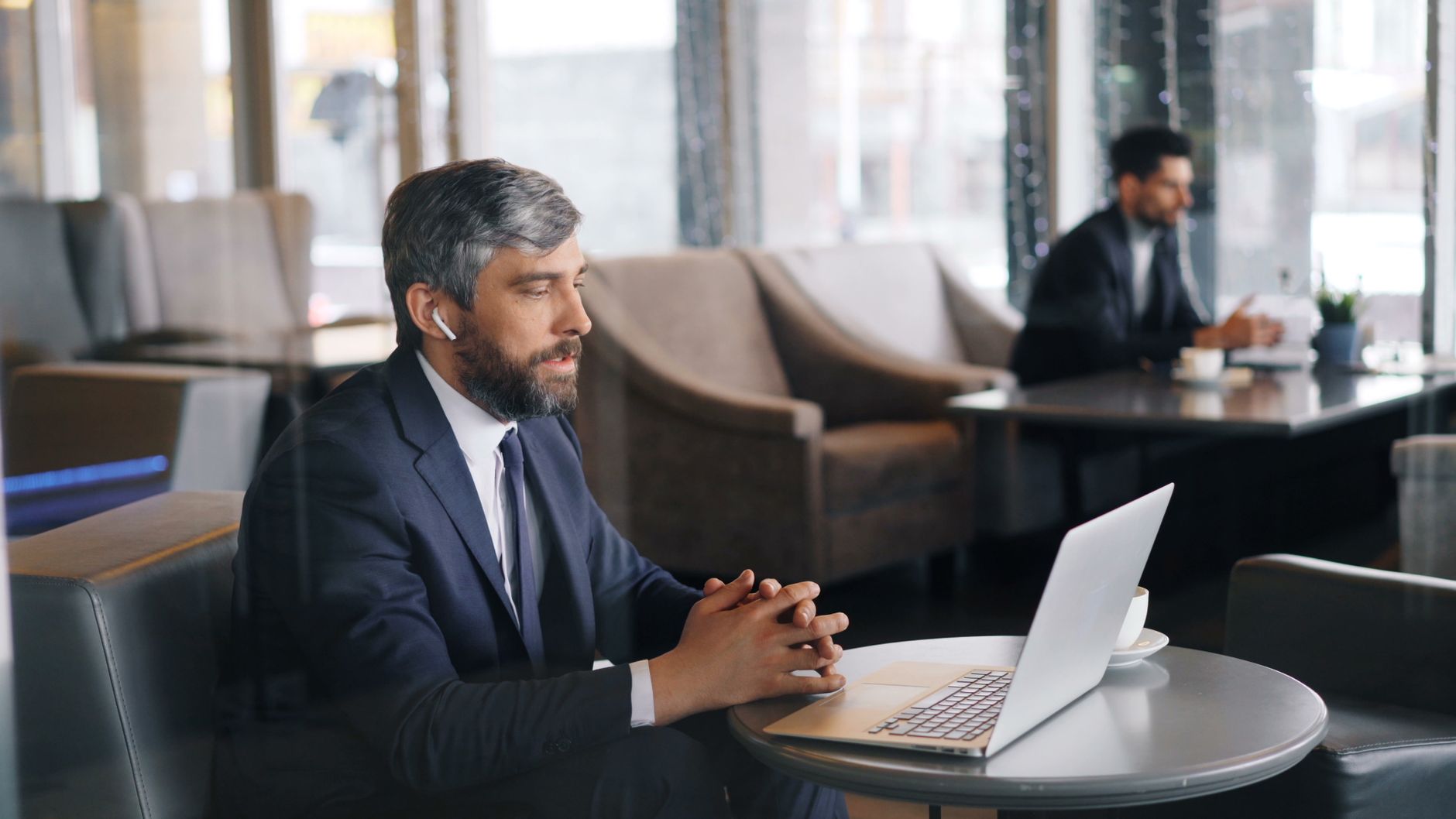 A man in a suit wearing a wireless earphone looks at a laptop on a café table during a video call.