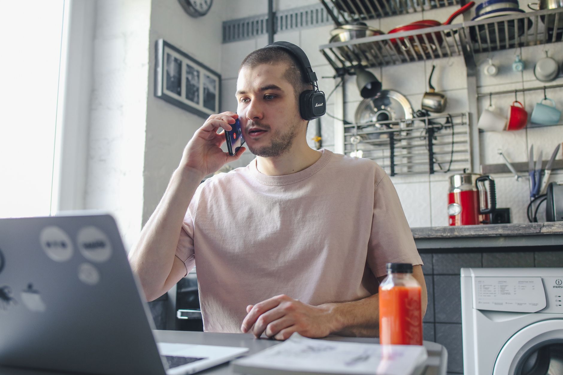 A man wearing headphones takes a phone call at a kitchen table, with a laptop open in front of him.